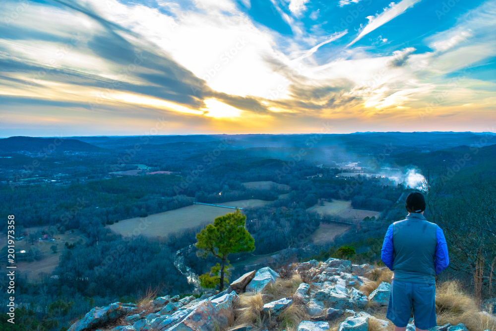 Man in a blue jacket standing on a rocky peak, overlooking a scenic valley during sunset with dramatic clouds.