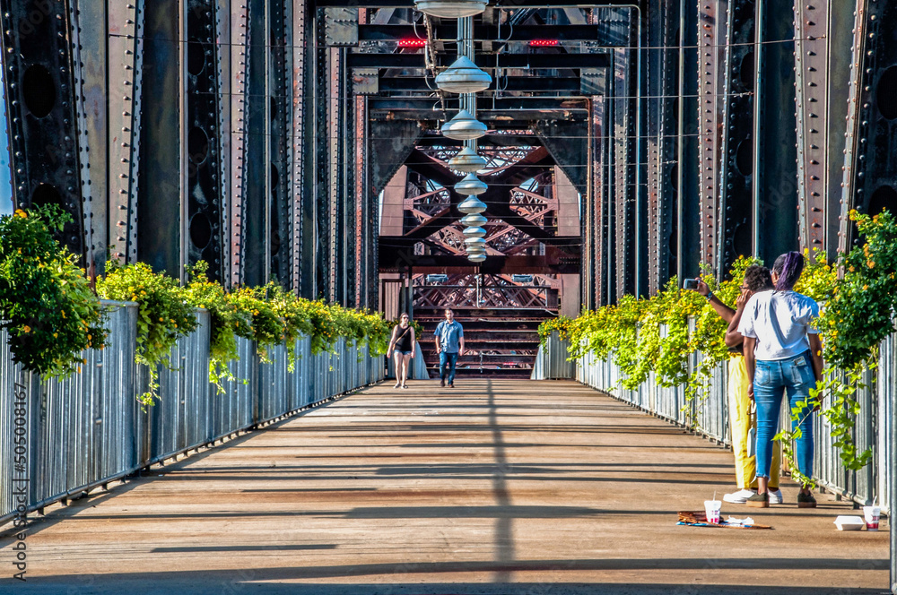People walking on an industrial bridge walkway adorned with green plants, steel beams, and overhead lamps.